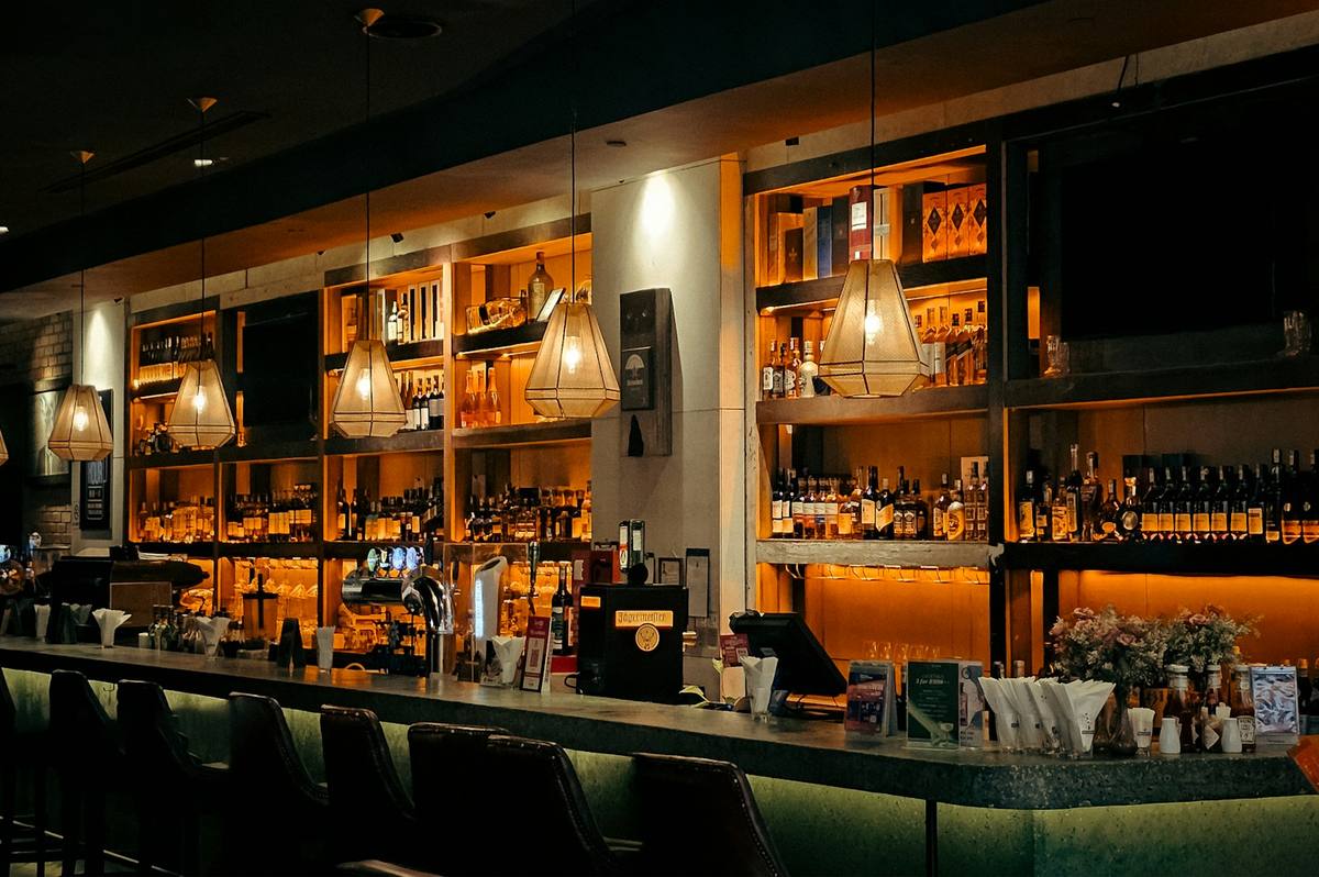 Warm, dimly lit bar counter with backlit shelves of bottles and bar stools